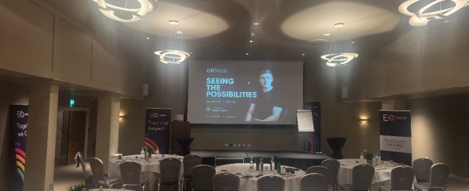 Conference room set up for a professional business event, featuring round tables, presentation screen, stage, and modern circular ceiling lights. A keynote slide titled “Seeing the Possibilities” is displayed at the front of the room. AI Keynote Speaker 2026.