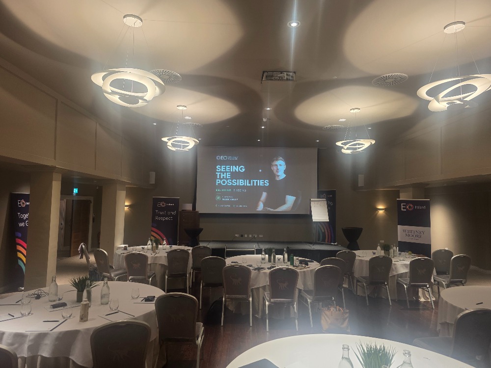 Conference room set up for a professional business event, featuring round tables, presentation screen, stage, and modern circular ceiling lights. A keynote slide titled “Seeing the Possibilities” is displayed at the front of the room. AI Keynote Speaker 2026.