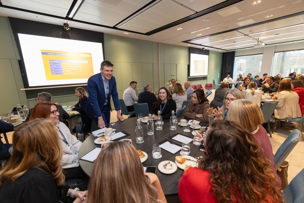Mark Kelly facilitating a group AI workshop, moving between tables of business leaders in a full conference room