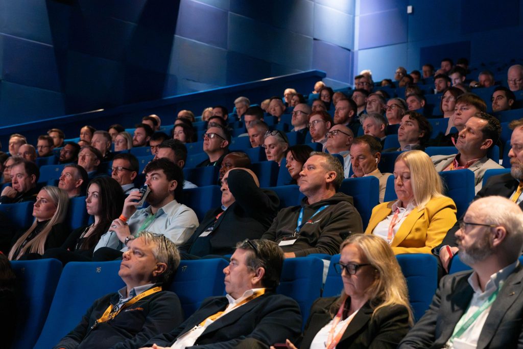 An audience seated in a dark blue auditorium, looking up toward the stage during Mark Kelly's AI presentation at the Arrow Conference.
